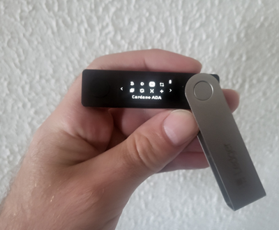 Hand holding Ledger Nano X hardware wallet displaying Cardano ADA app on the OLED screen.